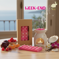 la photo présente une Tablette de fondants parfumés en cire d'olive naturelle, sans produits chimiques, vegan, fragrances fruitées et florales, emballée dans un étui avec fenêtre transparente pour voir le produit à l'intérieur. On voit également sur la photo une noix de coco fraiche, des gousses de vanille brillantes, des quartiers d'agrumes, des framboises et un brule parfum blanc duquel s'élèvent des effluves envoutantes. 