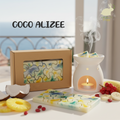 la photo présente une Tablette style puzzle de fondants parfumés en cire d'olive naturelle, sans produits chimiques, vegan, couleur verte, blan, bleu, jaune, emballée dans un étui couleur kraft avec fenêtre transparente pour voir le produit à l'intérieur. On voit également sur la photo des gousses de la noix de coco, de l'ananas, des groseilles et des banaes et un brule parfum blanc duquel s'élèvent des effluves envoutantes
